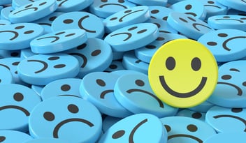 Pile of blue sad faces with single happy face in yellow